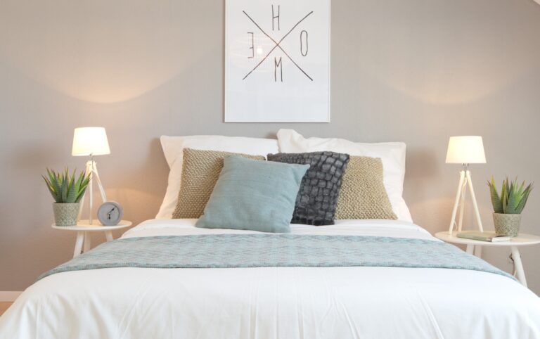 Home Staging für Erbimmobilie. Durch die defente taubenfarbige Wand im Hintergrund, davor ein Doppelbett mit weissen Lacken und weiss, beige, hellblau Kissen. Beidseitig Nachtische mit Lampe und kleine Aloepflanze.