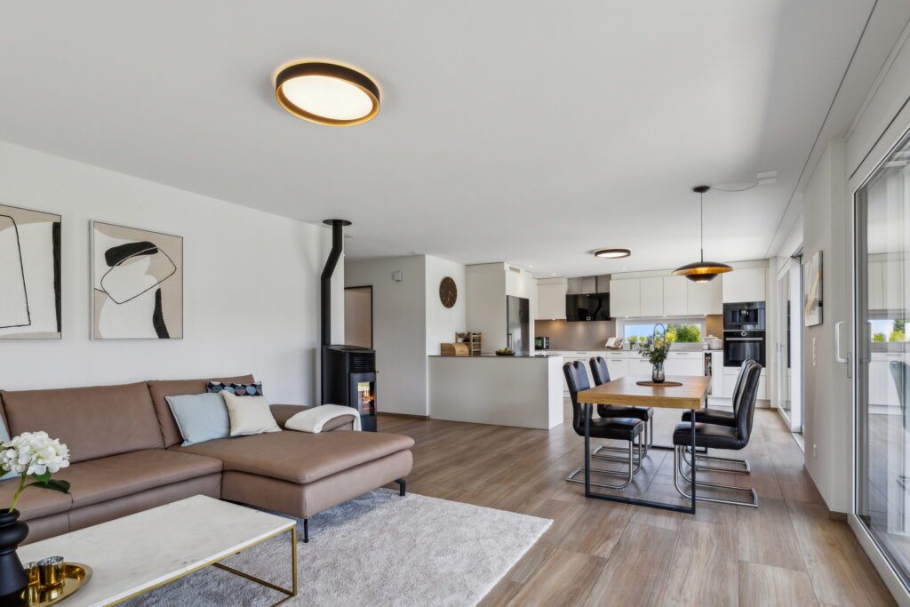 Bewohnte Immobilie, Wohn-, Essen-, Küchenbereich in Beige und braunem Farbton gehalten. Braunes Leder Ecksofa mit weissem Marmorclubtisch, schwarzer Kaminofen an der Wand. Brauner Esstisch in der Mitte mit schwarzen Lederstühlen und im Hintergrund die weisse offene Küche.