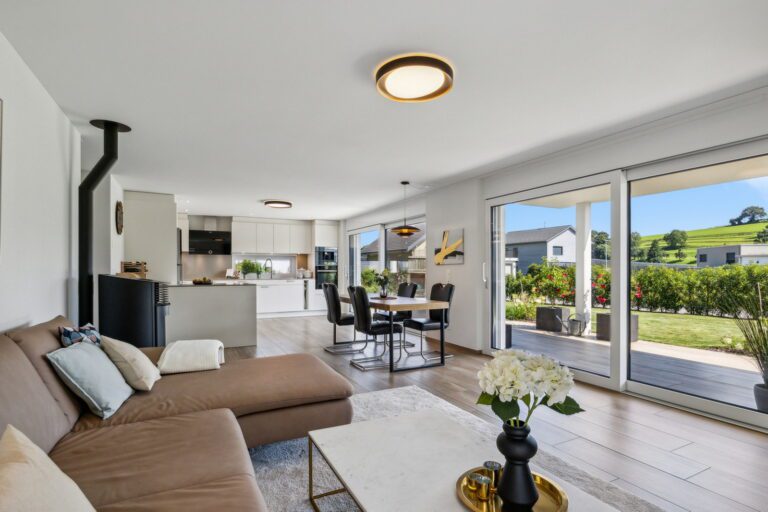 Home Staging für Bewohnte Immobilie, Wohn-, Essen-, Küchenbereich in Beige und braunem Farbton gehalten. Braunes Leder Ecksofa mit weissem Marmorclubtisch, schwarzer Kaminofen an der Wand. Brauner Esstisch in der Mitte mit schwarzen Lederstühlen und im Hintergrund die weisse offene Küche.